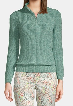 Frau in einem tealfarbenen gestrickten Zip-Up-Pullover und weißen Hosen mit kleinem, mehrfarbigem Blumenmuster, steht vor einem einfarbigen Hintergrund.