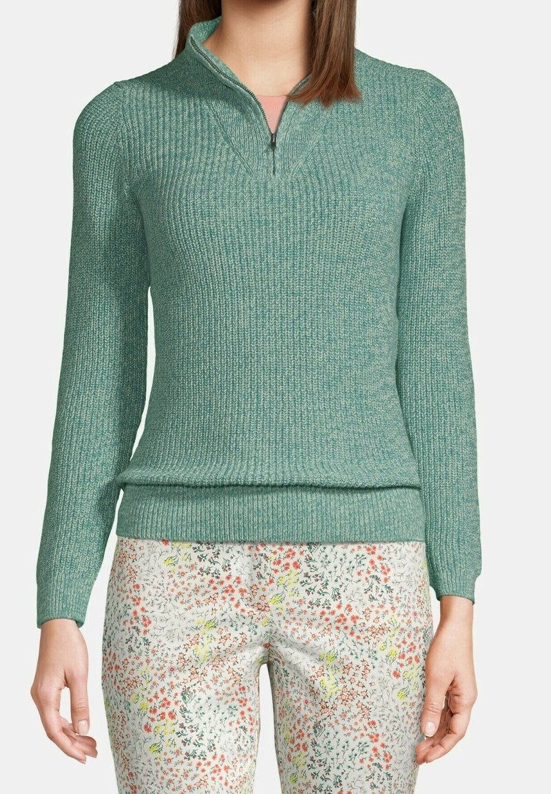 Donna che indossa un maglione a zip in maglia turchese e pantaloni bianchi con un piccolo motivo floreale multicolore, in piedi davanti a uno sfondo semplice.