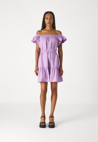 Robe violette à épaules dénudées avec des manches bouffantes et un corsage smocké. Dotée d'une taille ceinturée et d'un ourlet à volants superposés. Portée avec des chaussures à plateforme noires.