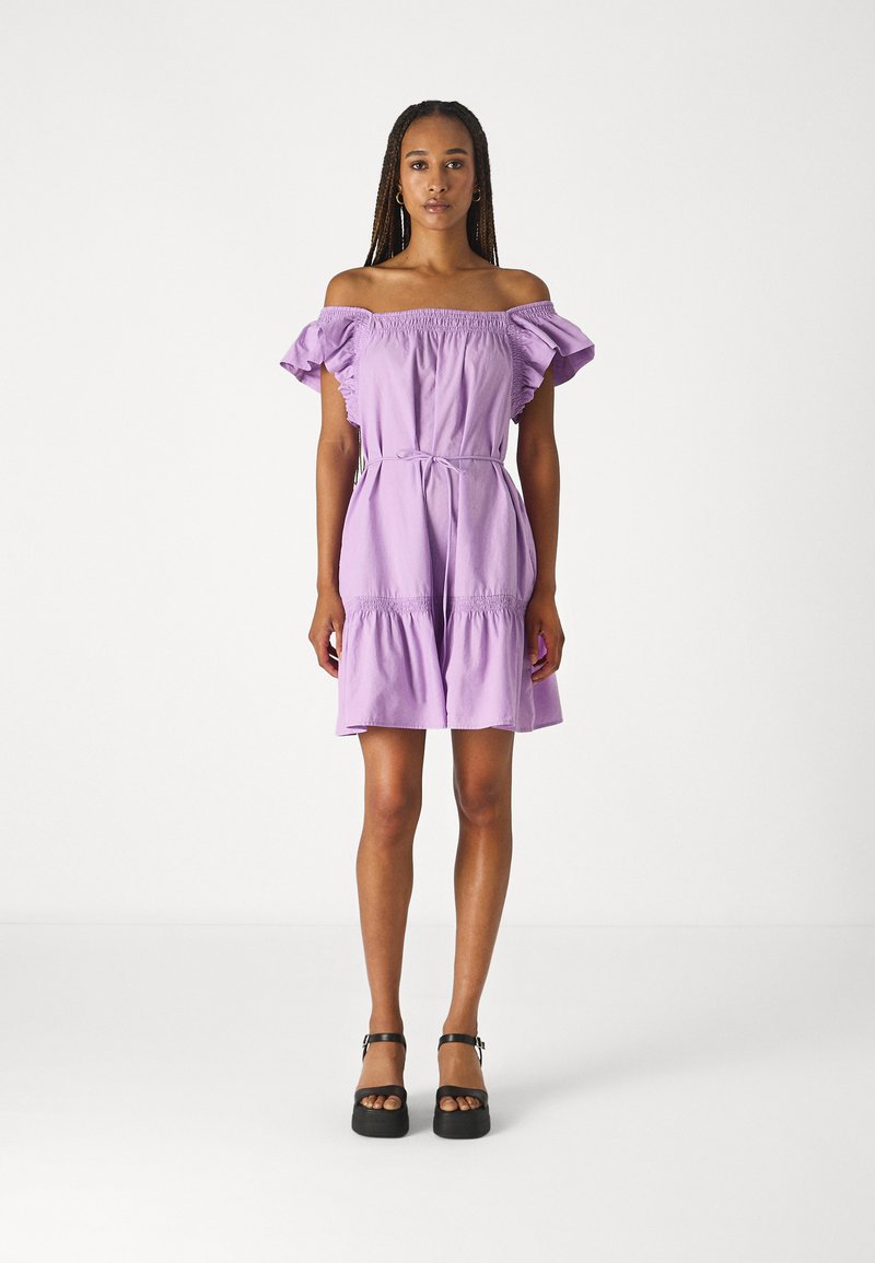 Robe violette à épaules dénudées avec des manches bouffantes et un corsage smocké. Dotée d'une taille ceinturée et d'un ourlet à volants superposés. Portée avec des chaussures à plateforme noires.