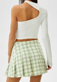 Kvinna som bär en vit enaxlad långärmad topp och en grön- och krämfärgad rutad plisserad minikjol, sedd bakifrån.