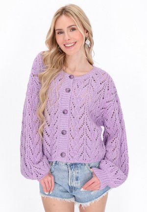Cardigan en tricot lavande avec un motif de feuilles, encolure ronde et gros boutons. Présente des manches ballon et un design cropped, porté par-dessus des shorts en denim.