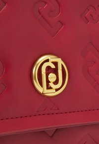 Borsa a tracolla in pelle rossa con motivo in rilievo, logo circolare in tono oro e dettagli cuciti lungo i bordi.