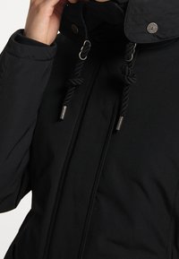 Giacca nera imbottita con colletto alto, accessori in metallo, coulisse e una texture liscia. Presenta un bottone sul colletto e chiusura con zip.