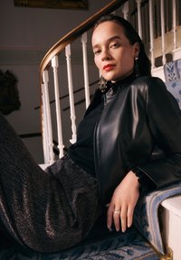 Schwarze Lederjacke, schwarzes Oberteil, funkelnde graue weite Hose, Rüschensaum, kräftiger roter Lippenstift und große, hängende Ohrringe. Weiche Texturen sind sichtbar.