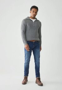 Grau gerippter Pullover mit halbem Reißverschluss, lange Ärmel, getragen über einem weißen Hemd, kombiniert mit blauen Slim-Fit-Jeans und braunen Stiefeln. Einfarbiger Hintergrund.