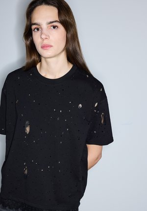 Młoda kobieta z długimi brązowymi włosami, ubrana w czarną, postrzępioną koszulkę z krótkim rękawem, stoi na tle jednolitego, jasnoszarego tła.