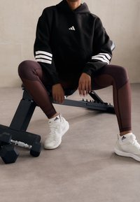 Czarna bluza z kapturem w białe paski, bordowe legginsy z kontrastowymi panelami i białe sneakersy z czarnymi akcentami. W pobliżu ciężka hantla.