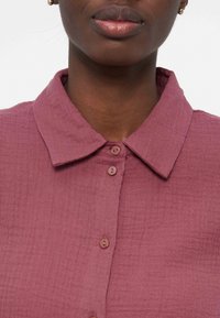 Artimas vaizdas asmens, dėvinčio struktūruotą rausvai violetinę su sagutėmis marškinių palaidinę su apykakle ir mažais apvaliais mygtukais, dalinai matomos lūpos ir auskaras.