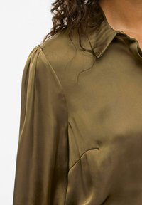 Blusa satinata verde oliva con colletto e maniche a palloncino. La texture liscia e la leggera lucentezza ne esaltano l'aspetto elegante.