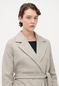 Giovane con capelli ricci corti che indossa un cappotto di lana beige con cintura sopra una maglietta nera, su uno sfondo chiaro uniforme.
