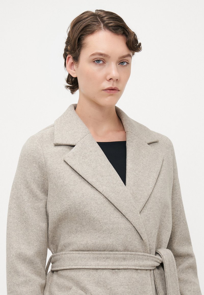 Jeune personne avec des cheveux bouclés courts portant un manteau en laine beige ceinturé sur un haut noir, devant un fond clair uni.