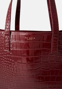 Saco tote bordô com textura de crocodilo, acabamento suave e brilhante, duas alças robustas e um rótulo de logótipo dourado.