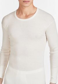 Maglietta termica a maniche lunghe di colore crema con una texture a coste, scollo tondo e design aderente, adatta per essere indossata a strati.