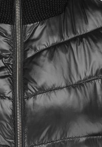 Schwarze Pufferjacke mit glänzender Oberfläche, gestepptem Muster und einem gerippten schwarzen Strickkragen. Zentrale Reißverschlussöffnung.