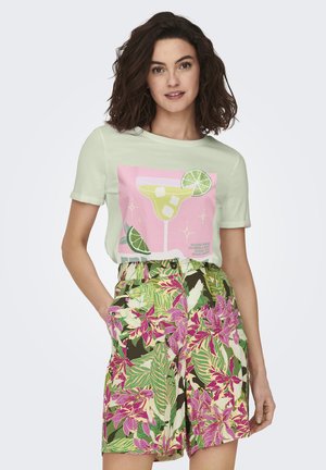 Mujer con camiseta verde pálido con gráfico de copa de cóctel y pantalones cortos con estampado floral tropical, de pie con una mano en el bolsillo.