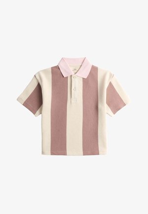 Ung dreng med krøllet hår iført en beige og lyserød stribet kortærmet polo-shirt og lyseblå jeans med hænderne i lommerne.