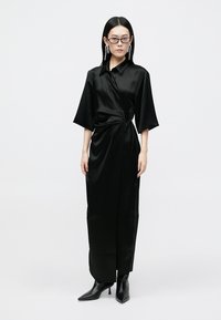 Robe en satin noire avec un design portefeuille, manches courtes et taille ceinturée. Associée à des bottines noires pointues et des boucles d'oreilles distinctives.
