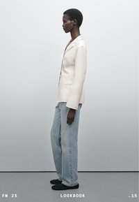 Béžový blazer s klopami, doplněný o světle modré rovné džíny. Černé boty. Osoba je zobrazena v profilu na neutrálním pozadí.