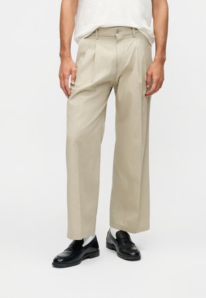 Mies, jolla on beigejä poimuttuja leveälahkeisia housuja, valkoiset sukat, mustat nahkakengät ja valkoinen lyhythihainen paita, seisoo yksinkertaisella taustalla.