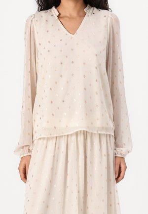 Kobieta ubrana w kremowy zestaw bluzki z długim rękawem i spódnicy w metaliczne kropki, z falbankowym dekoltem w kształcie litery V, stojąca z ramionami swobodnie opuszczonymi wzdłuż boków.