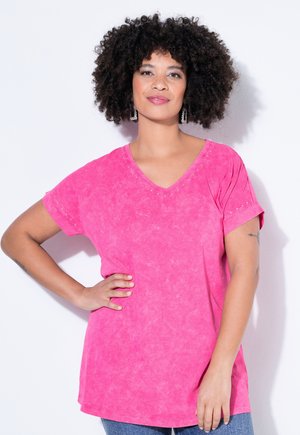 Kiharatukkainen nainen, joka pukeutunut kirkkaan pinkkiin V-kaula-aukkoiseen lyhythihaiseen paitaan, seisoo käsi lanteilla valkoista taustaa vasten.
