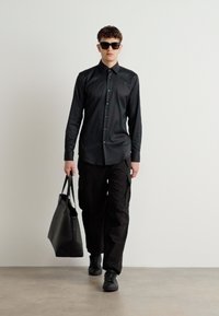 Camisa negra de botones, pantalones cargo y zapatillas negras. Lleva un grande bolso tote negro estructurado. Las gafas de sol completan el look.