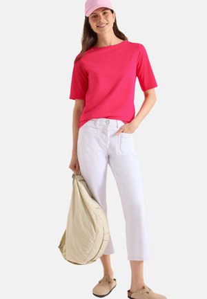 Kobieta uśmiechająca się w różowej czapce i koszuli, białe spodnie 7/8, brązowe wsuwane buty, trzyma dużą beżową torbę przy boku.