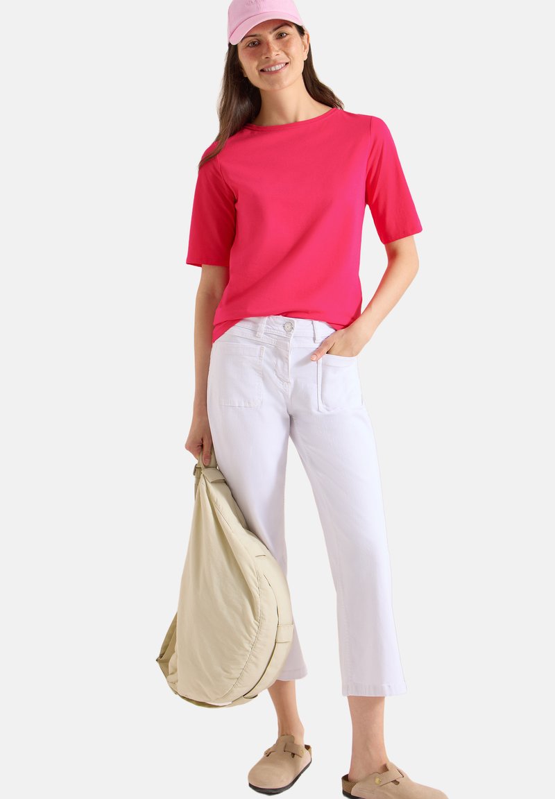 Femme souriante avec une casquette et une chemise roses, pantalon blanc court, chaussures beiges sans lacets, tenant un grand sac beige à ses côtés.