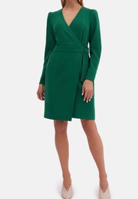 Robe verte enveloppante avec des manches longues, décolleté en V et ceinture à la taille. Tissu fluide, longueur genoux, avec un léger drapé et une silhouette structurée.
