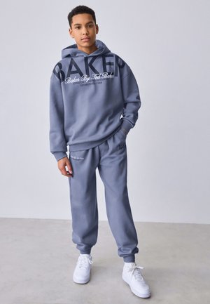 Teismeline, kes kannab sinist "Baker by Ted Baker" huppi ja sobivaid spordipükse, seisab ühe käe taskus ja valgete tossudega hallil põrandal.