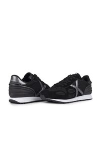 Zapatillas negras con parte superior de malla y acentos de gamuza. Cuenta con un diseño de rayas plateadas y una suela de goma negra. Estética minimalista.