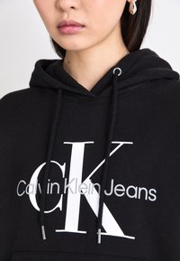 Černá bavlněná mikina s kapucí, na které je velké bílé logo "CK" s šedým nápisem "Calvin Klein Jeans". Obsahuje nastavitelný šňůrkový tah.