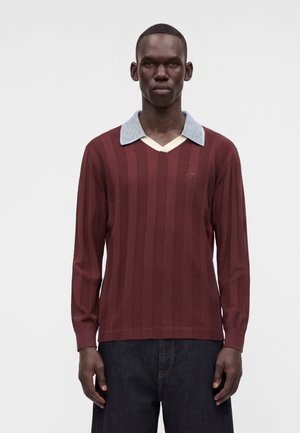 Homme portant un pull en tricot bordeaux à manches longues avec des rayures verticales et un col bleu clair, debout devant un fond uni.