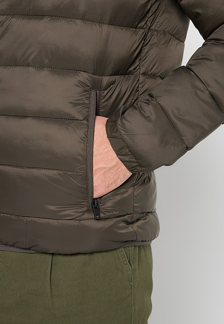 Chaqueta acolchada verde oliva con un bolsillo lateral con cremallera, que presenta una textura suave y brillante, y un diseño contorneado.