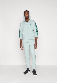 Gai piparmētru zaļa kapuce un sporta bikses ar zaļām sānu svītrām. Materiāls izskatās mīksts; ar rāvējslēdzēju un kapuci. Melni sporta apavi.