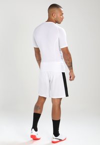 Hvit atletisk skjorte og shorts, tettsittende design, med svarte sidestreker på shortsen, iført svarte sokker og hvite sneakers. Glatt tekstur.