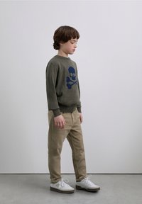 Olive green sweater with a navy skull and crossbones design, paired with beige pants and white sneakers, standing on a smooth surface.