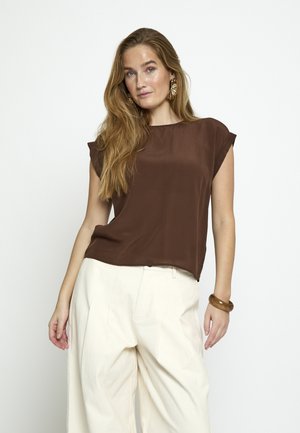 Pitkähiuksinen nainen, jolla on suuret kultaiset korvakorut, ruskea lyhythihainen pusero, leveät valkoiset housut ja ruskea rannekoru, seisoo.