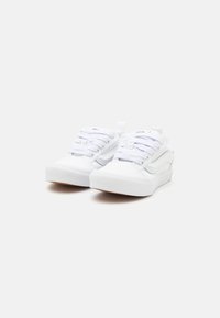 Un paio di sneakers bianche basse con suole spesse e lacci bianchi su uno sfondo chiaro e uniforme.
