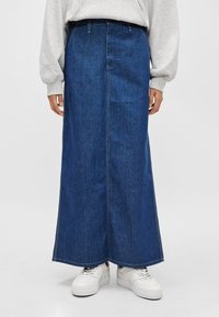 Saia maxi em denim, azul escuro com costuras cosidas, cintura alta, silhueta reta, combinada com ténis brancos. Tecido liso e durável.