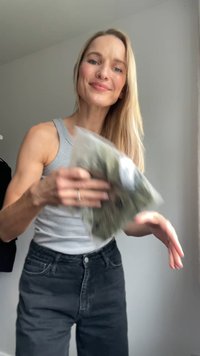 En person i en grå linne håller en genomskinlig påse med gröna föremål. De bär högmidjade svarta jeans. Bakgrunden är neutral.