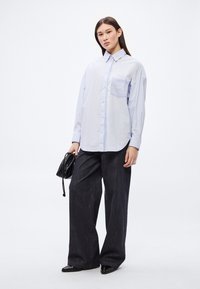 Svetlomodrá košeľa na gombíky s jedným náprsným vreckom, v kombinácii s tmavými džínsami s širokými nohavicami a čiernymi lodičkami na podpätku. V ruke matná čierna kabelka.