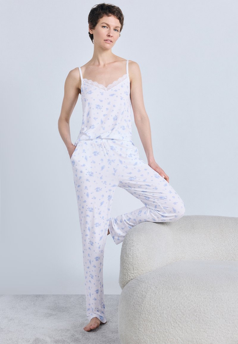 Γυναίκα που στέκεται ξυπόλητη με το ένα πόδι πάνω σε μια υφασμάτινη, κυρτή λευκή καναπέ, φορώντας λευκή πιτζάμα με ανοιχτό μπλε λουλουδάτα μοτίβα.