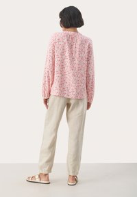 Långärmad rosa blus med bladmönster, samlad halsringning och lös passform. Matchad med ljusbeiga byxor och avslappnade sandaler synliga.