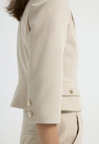 Beige přizpůsobený blazer s rukávy do čtvrtiny, který má na manžetách dva ozdobné zlaté knoflíky a design se spodničky.