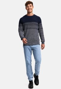 Navy- und graustrickpullover mit geripptem Design, kombiniert mit hellblauen Jeans und schwarzen Schnürstiefeln. Das Model steht vor einem neutralen Hintergrund.