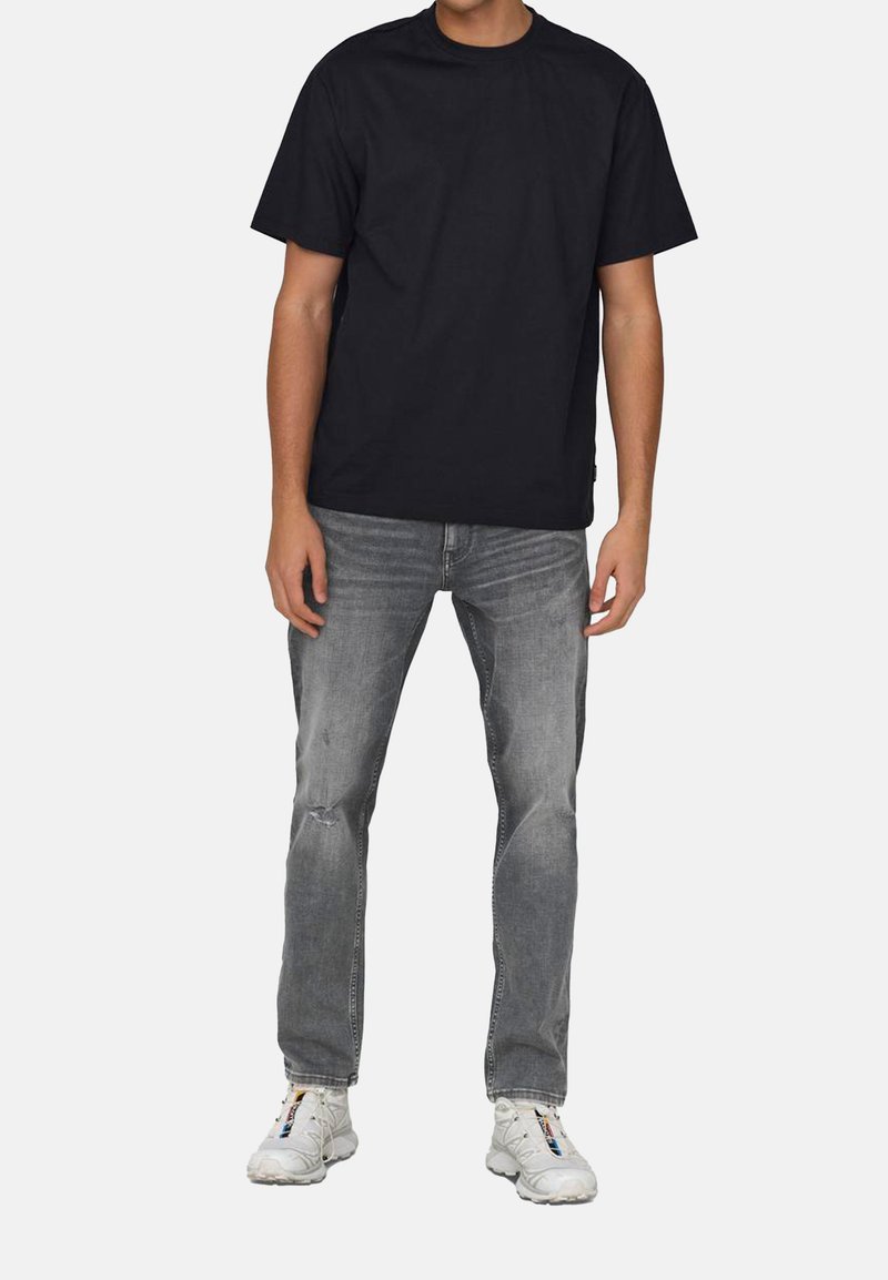 Schwarzes Baumwoll-T-Shirt mit Rundhalsausschnitt, kombiniert mit grauen, beschädigten Jeans. Die Schuhe haben ein mehrfarbiges Design mit weißen Akzenten.
