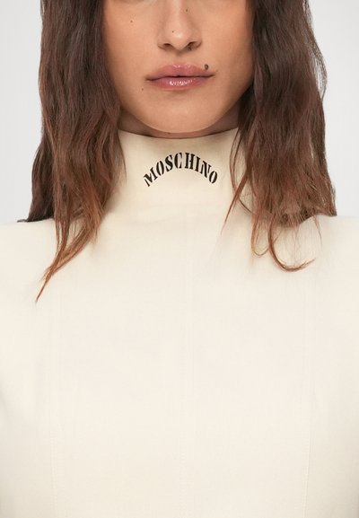 Ženska z dolgimi rjavimi lasmi, oblečena v belo majico z visokim ovratnikom, na katerem je natisnjeno "MOSCHINO", ter z majhno lepotno znamenje nad ustnicami.