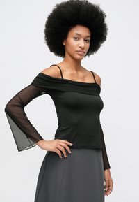 Femeie cu un afro mare și ondulat, purtând un top negru cu umeri goi, mâneci lungi din material transparent și fustă gri închis, pozând cu mâna pe șold.